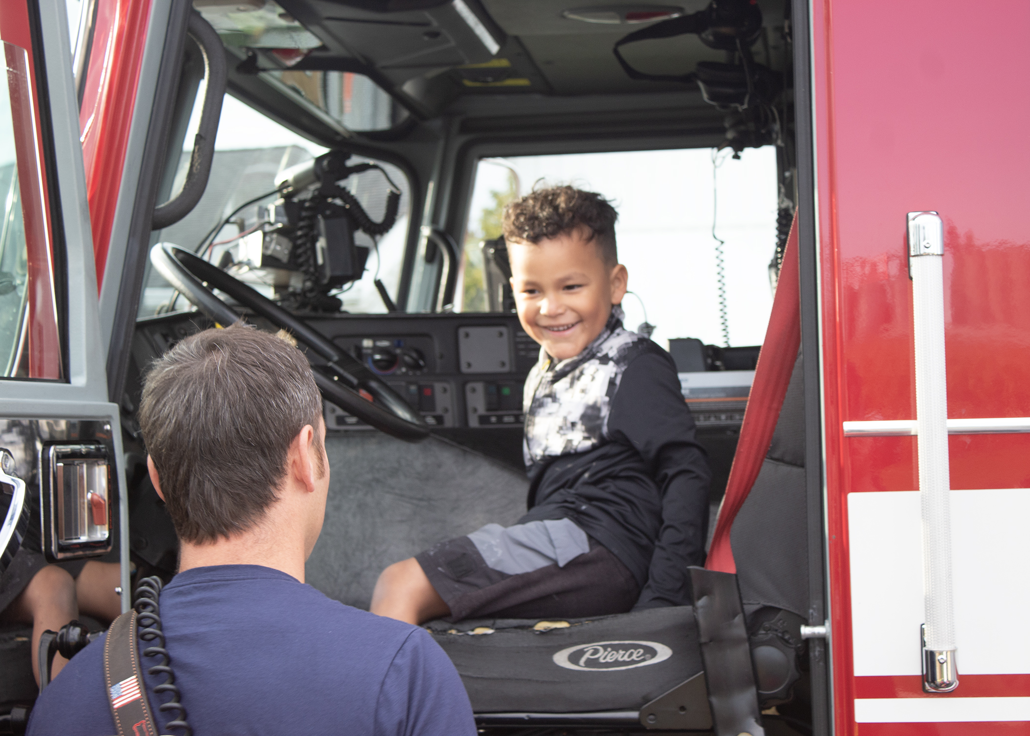 Smiling child sits in the front seat of a fire truck as a firefighter stands alongside the truck.