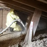 A GLCAP weatherization crew member in yellow Tyvek suit and face mask blows insulation into an attic. A ruler can be seen in the foreground, which measures the depth of the insulation as it is blown into the attic.