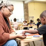 A woman teacher helps a small boy dip marbles into paint to create marble art while in a Head Start classroom.